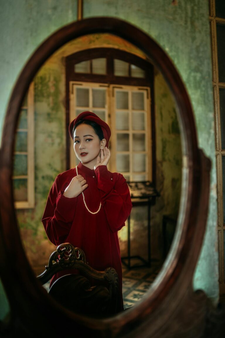 A woman in a red dress stands by an ornate mirror, embodying classic elegance indoors.
